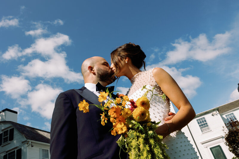Bride and groom kissing with blue skies and bright yellow and green florals at new england september wedding