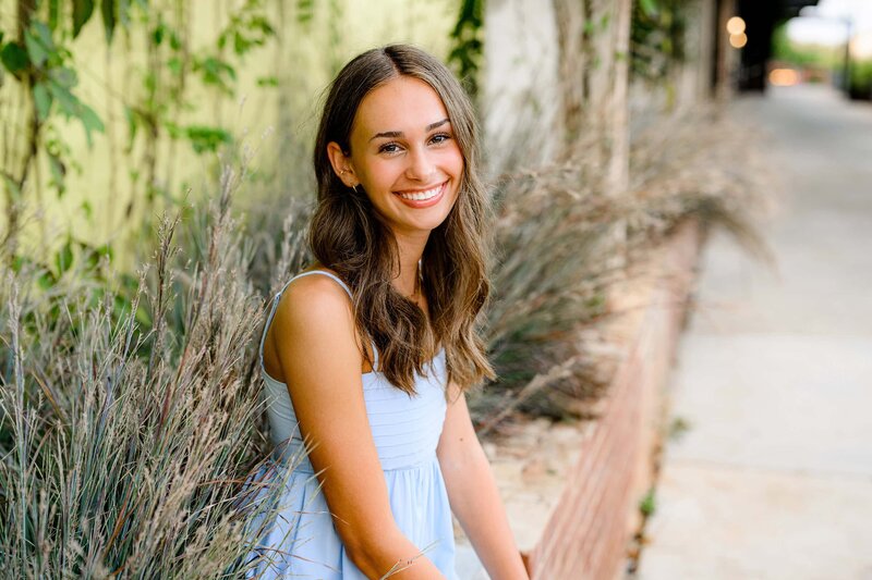dark hair grad sits on wall wearing a blue dress smiling