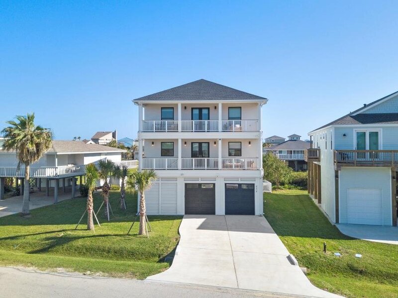 Straight-on exterior view of a newly built three-story coastal home with white siding, stacked front balconies, and two dark garage doors in Pirates Beach, Galveston, Texas.
