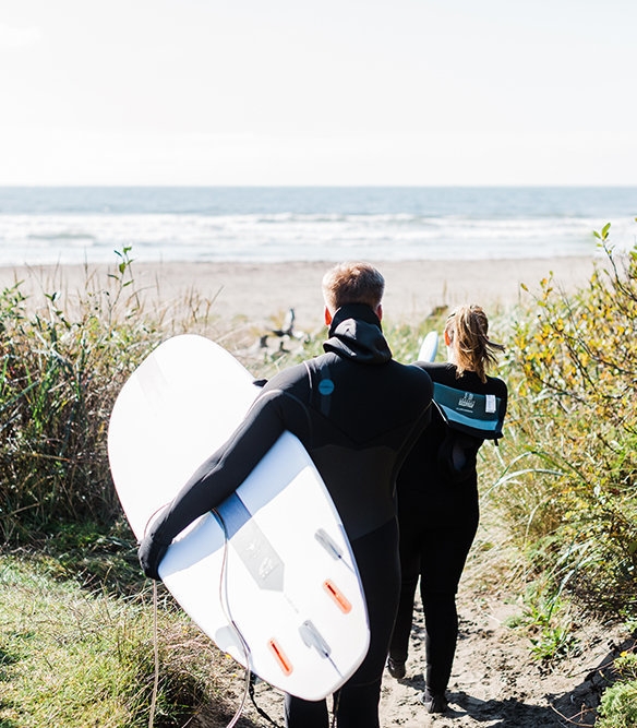 a couple heads out to take a surf together during their adventure engagement session on the washington coast with adventure elopement phototgrapher amy galbraith