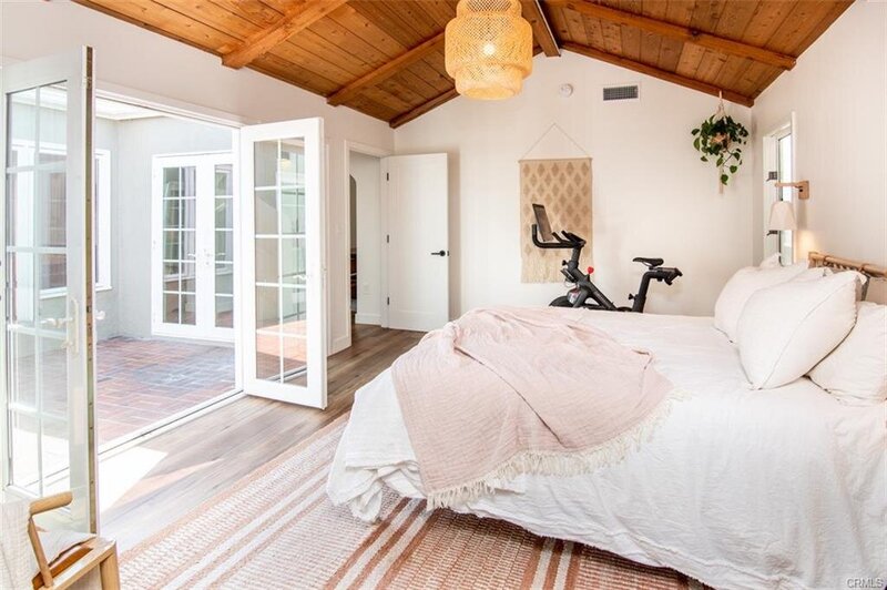 Boho bedroom with vaulted ceiling, bright walls, rattan chandelier, patio entryway, cozy furniture, and a Peloton bike in the corner.