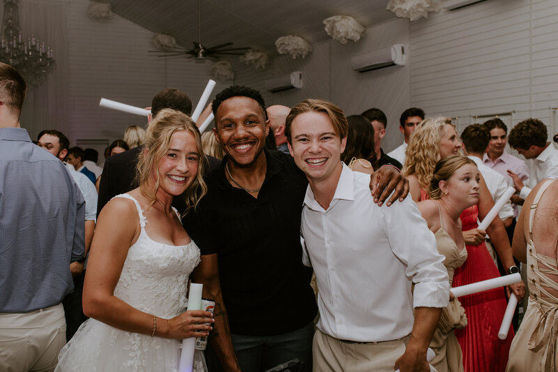 Bride and groom smiling with a guest during lively wedding reception, surrounded by friends and joyful celebration