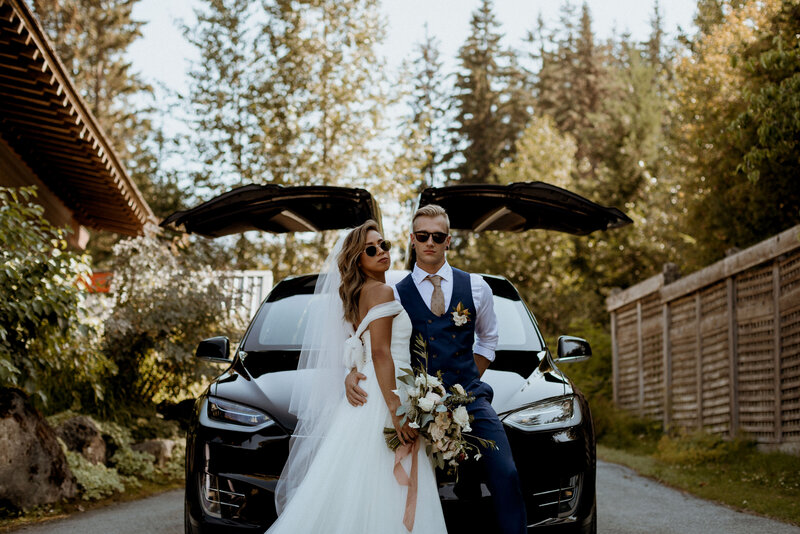 Chelsea and James standing in front of a Tesla at their wedding in Whistler