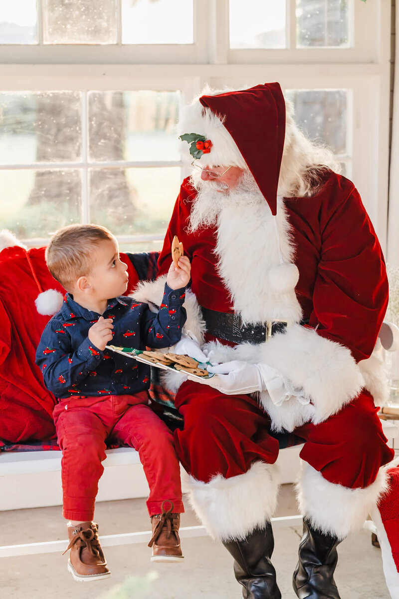 A young boy, in red pants and a Christmas truck shirt holds a cookie up for Santa. Santa is pretending to take a bite.