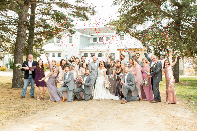 A wedding party and additional guests are standing closely together, throwing colorful confetti