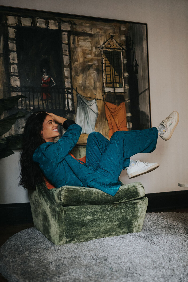 A brand designer reclines and laughs on a green armchair, wearing a teal outfit and white shoes. The cozy setup on the circular rug is completed by a large painting in the background, creating an inviting and stylish scene.