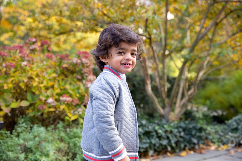 little boy looks at camera with falll foliage in the background at family photo shoot in Central Park Conservatory Gardens in NYC