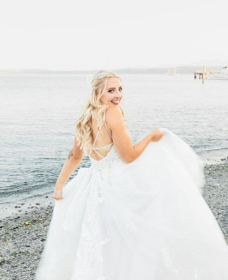 A bride turning with a radiant smile, holding her gown on a pebbled beach