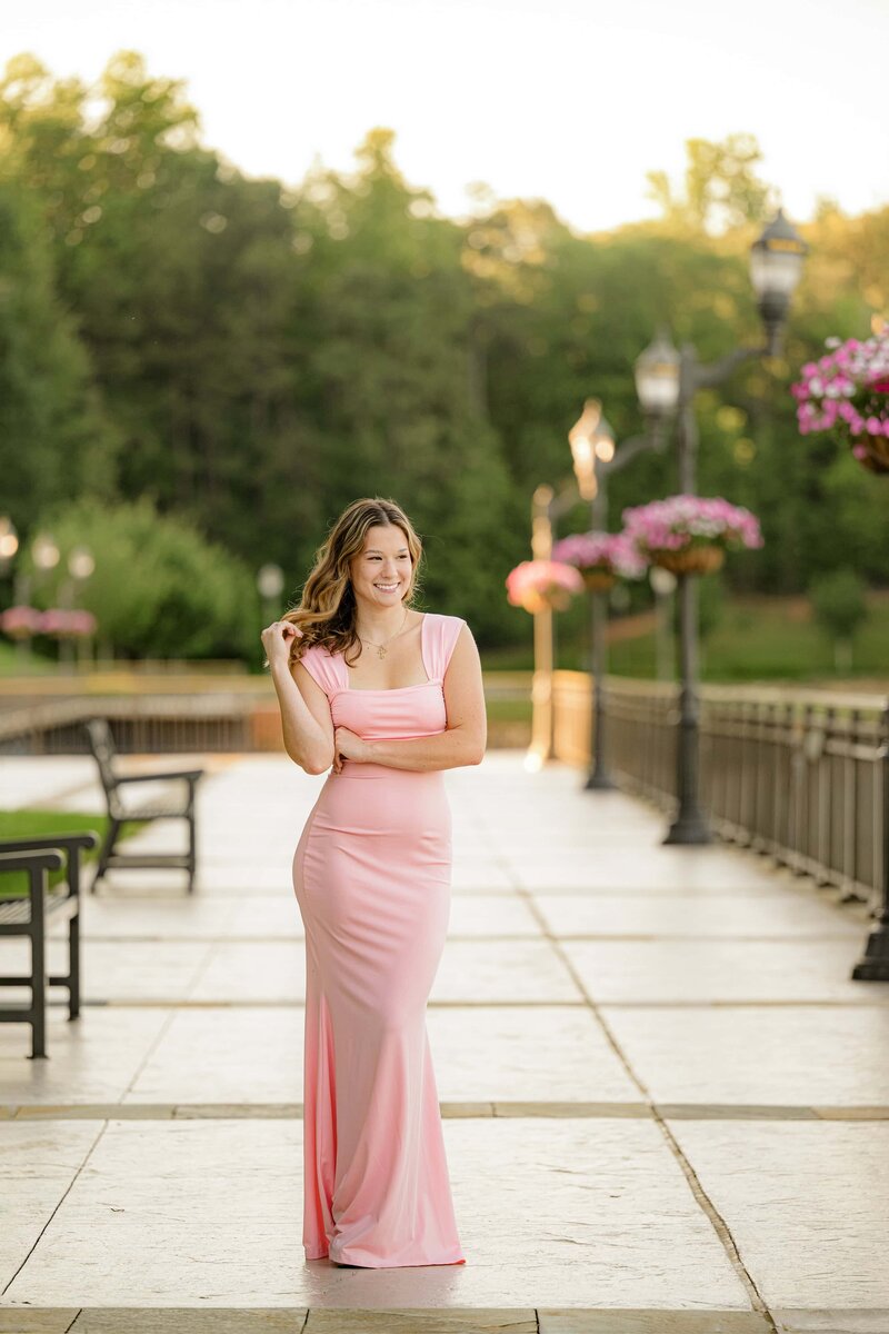 dark hair graduate stands in silk pink dress  with her hand in her hair looking off camera