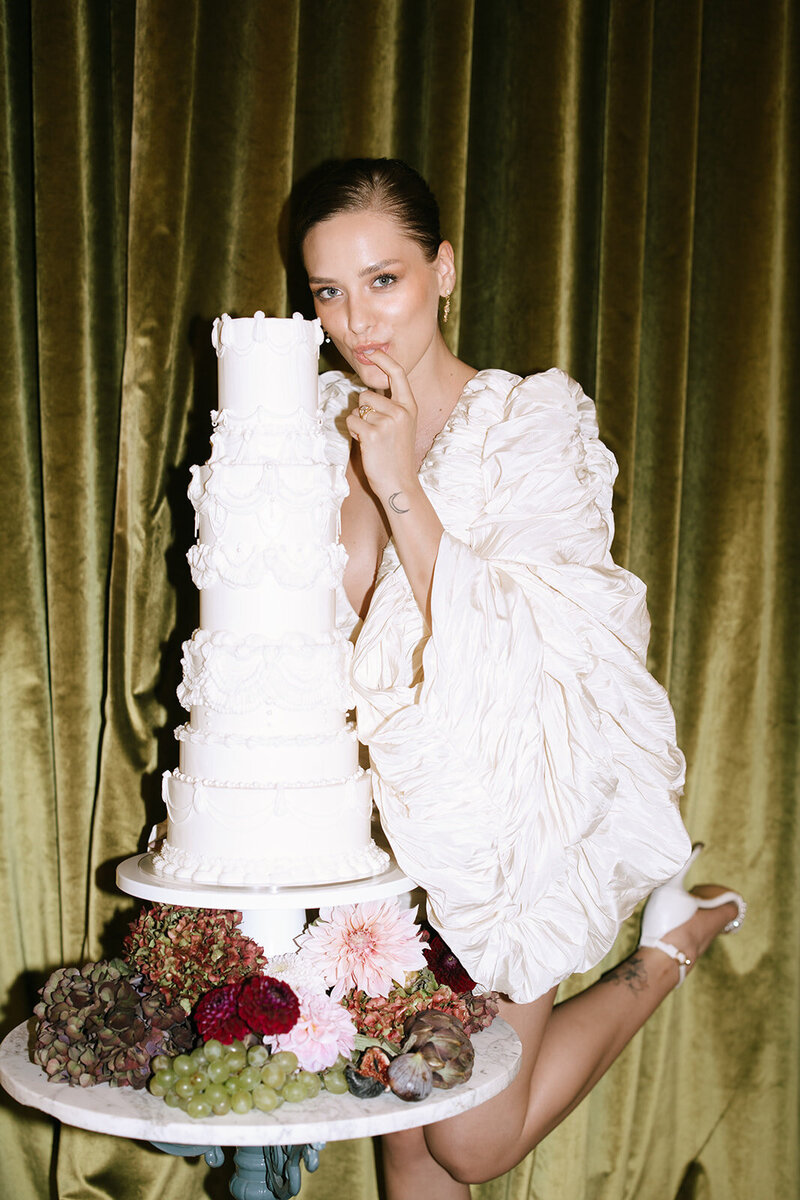 A bride playfully poses beside a tiered white wedding cake adorned with fresh flowers and fruit.