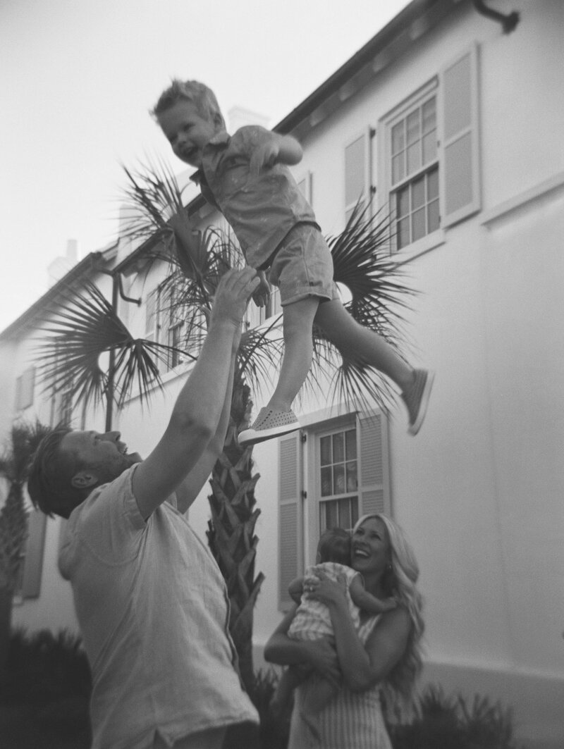 Black and white image of dad tossing son in the air while mom holding baby smiles at them