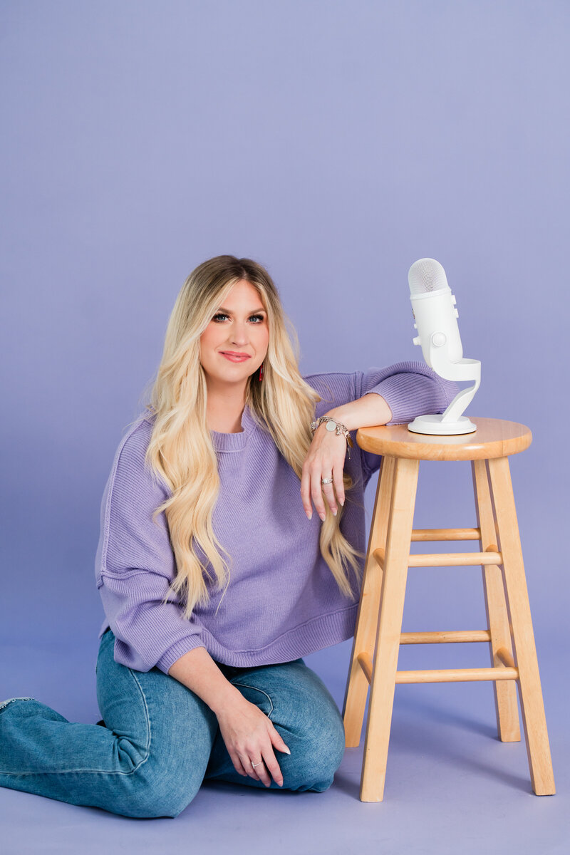 A headshot of Brittany Kues, podcast coach, sitting on the floor with her arm draped over a stool with a podcast mic sitting on top.