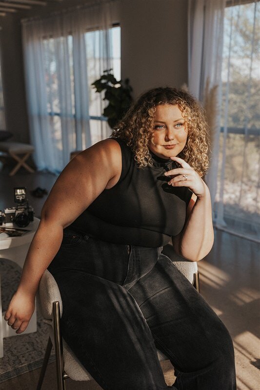 Andrea Cakmar sits thoughtfully on a chair in a well-lit room, with her elbow resting on her knee and hand touching her chin. She is wearing a black sleeveless top and black jeans, with a camera on a nearby table.