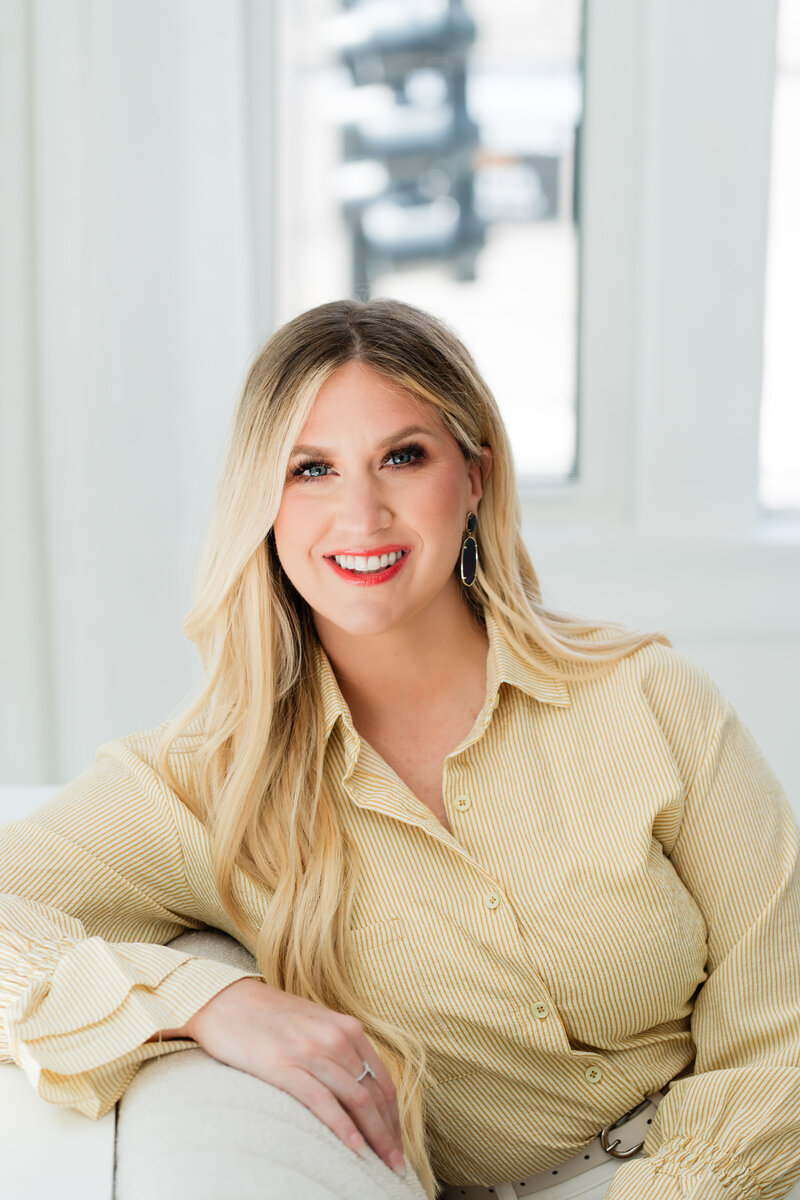 A headshot of Brittany Kues, podcast coach, smiling at the camera and sitting on a couch with her arm casually draped over the arm.
