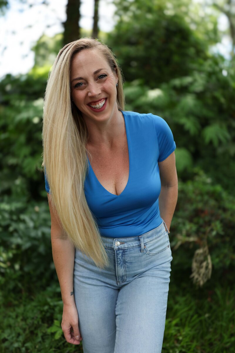 Smiling woman wearing blue top, standing outdoors in front of lush green trees in the background.