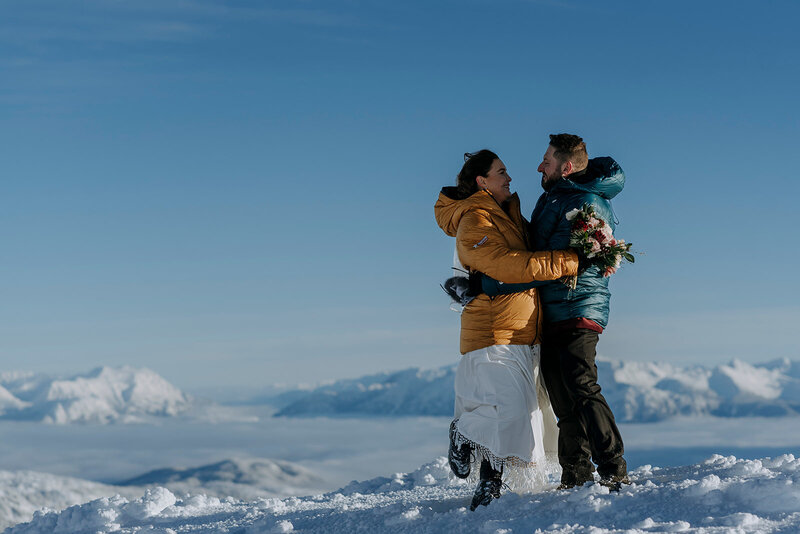 A bride and groom wearing a yellow and green puffy jacket hold each other on top of Rainbow Glacier in Whistler. There is snow all around and blue skies.