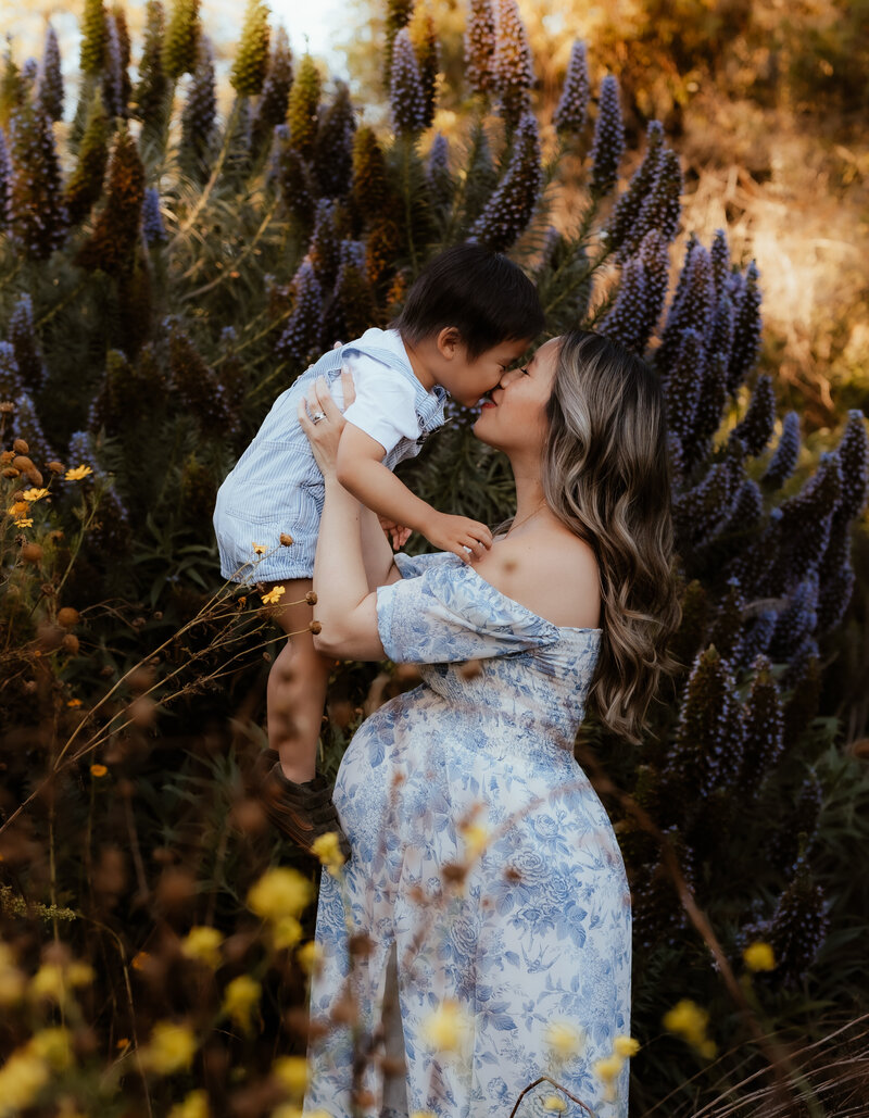 A pregnant mother in a gorgeous floral dress holds her toddler boy up in the air and rubs noses with him. 