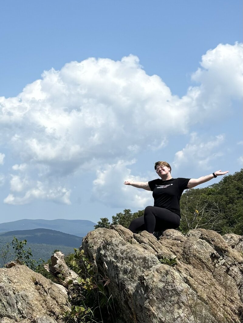 A Virginia intimate wedding photographer sits on top of a rock in Shenandoah National Park. She is wearing a black t-shirt that reads "it's always spicy in Appalachia". Her arms are stretched out to either side as she closes her eyes and breathes in the fresh air.