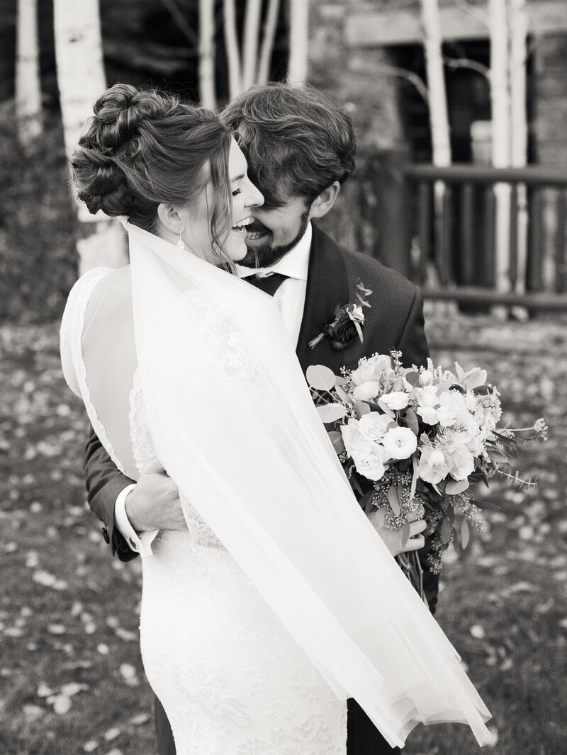 Black and white photo of a bride and groom hugging and laughing. The brides back is towards the camera and you can see her open back lace dress, and long flowing veil.