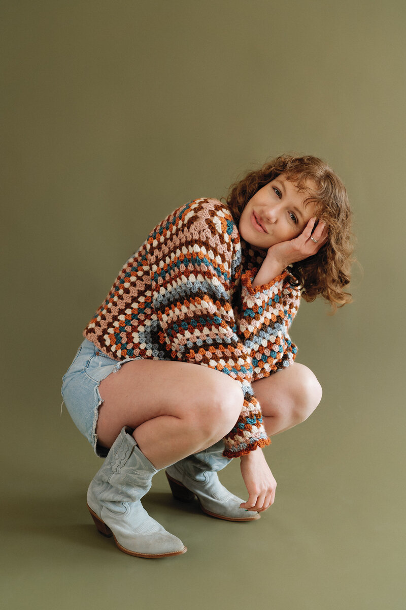 photographer wearing a colorful knitted sweater and blue cowboy boots and crouched down with a tilted head and smiling