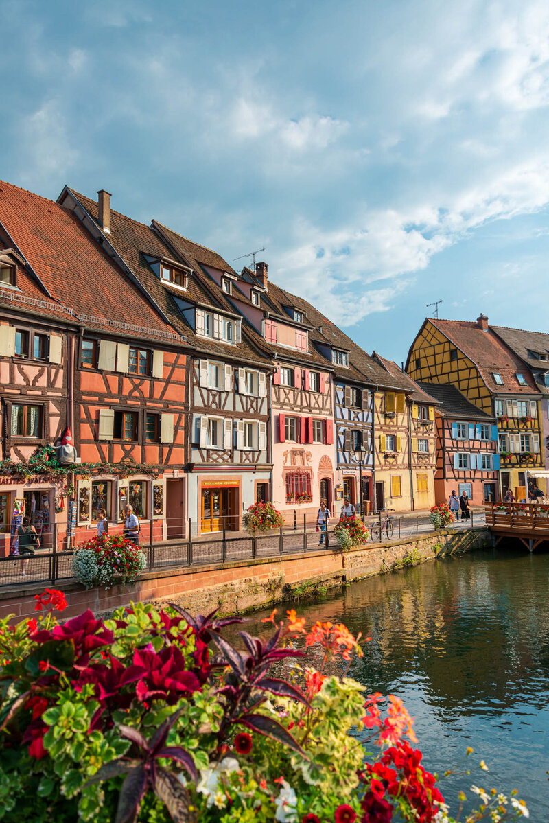 Colorful half-timbered houses lining a canal in a picturesque European town, with vibrant flowers in the foreground and people walking along the waterfront under a partly cloudy sky.