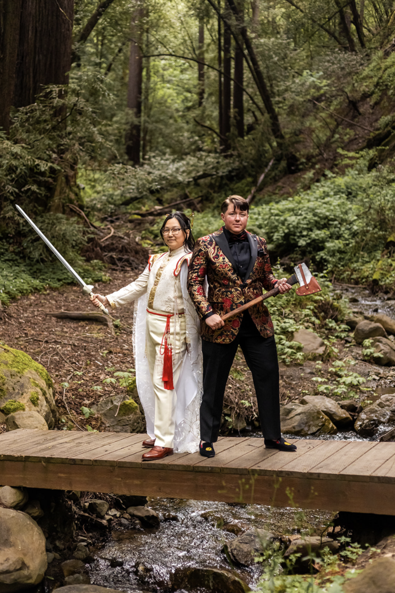 N nonbinary couple stand on a bridge in the woods in elaborate outfits. One holds a sword, the other an axe.