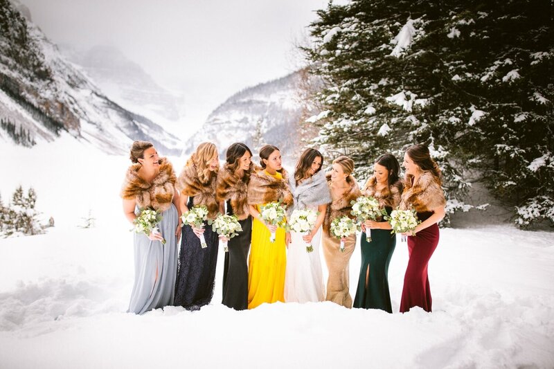 Bridesmaids share a joyful moment outside at Lake Louise during the winter months, with a snow-covered frozen lake and the stunning Victoria Glacier as the backdrop.