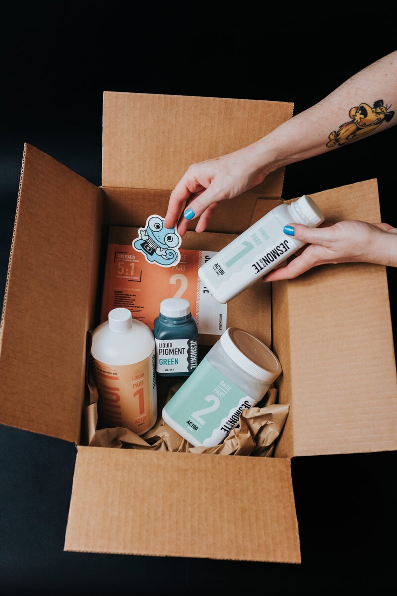 Two hands reach inside of a cardboard box with multiple paint products.