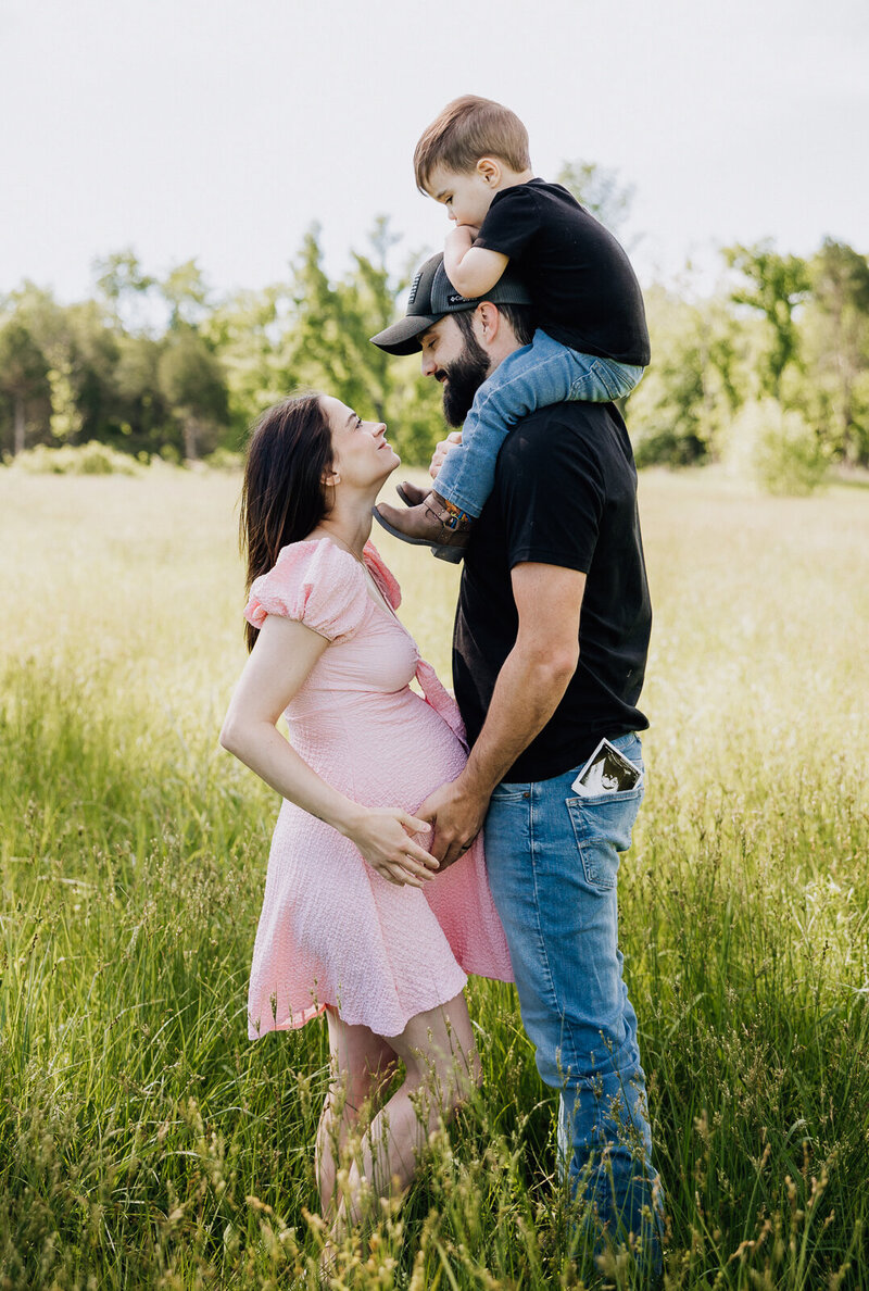Family maternity session in a meadow, parents holding mother's belly with their child on dad's shoulders.
