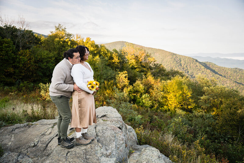 A groom holds onto his bride's hips and places his chin on her shoulder as the two look out at the view at Timber Hollow Overlook at Shenandoah National Park in Virginia on their elopement day.