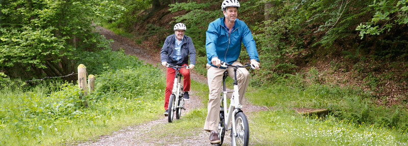 Two adults riding bicycles on a forest trail, wearing helmets and casual outdoor clothing, surrounded by lush green trees and foliage.