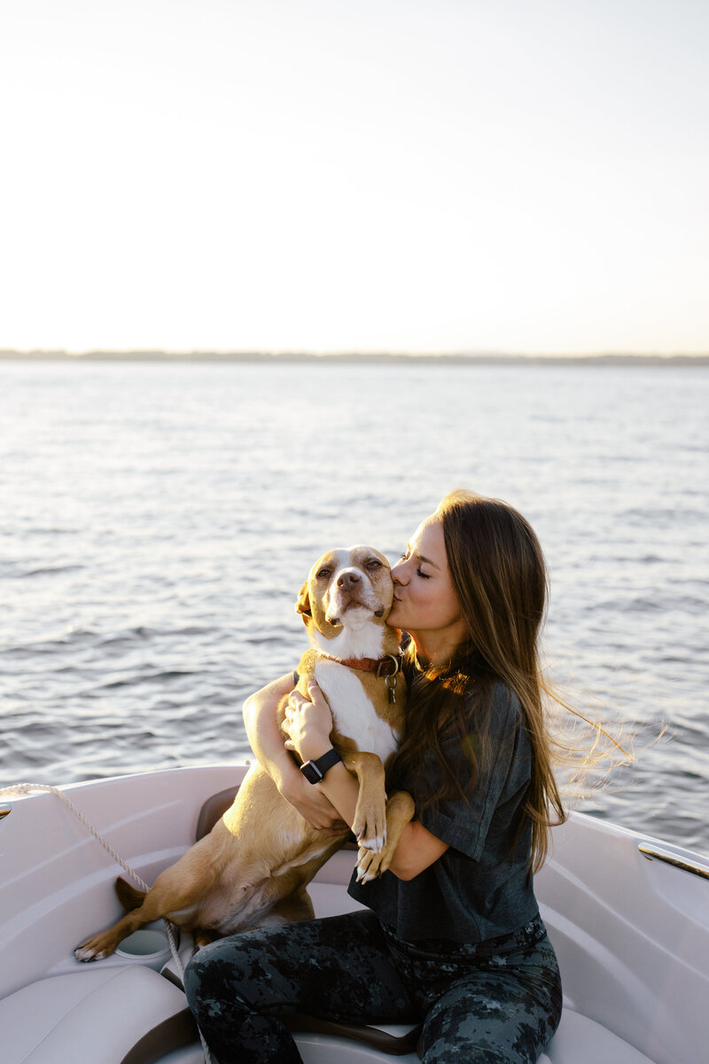 Girl with brown hair wearing a grey shirt is holding a tan and white dog on a boat with lake water in background by Charleston wedding photographer, Stephanie Bailey Photography.