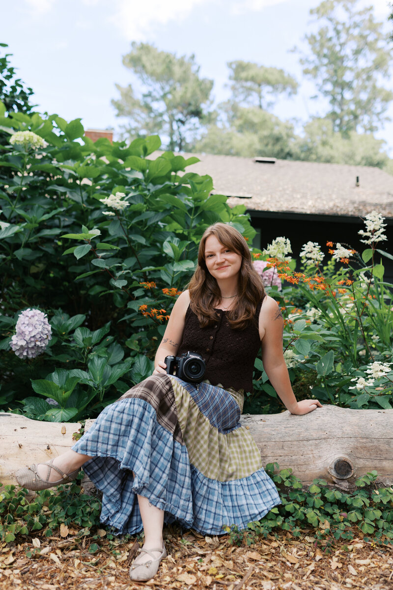 Photographer sitting on a log with her legs crossed in a colorful skirt and brown shirt looking at the camer
