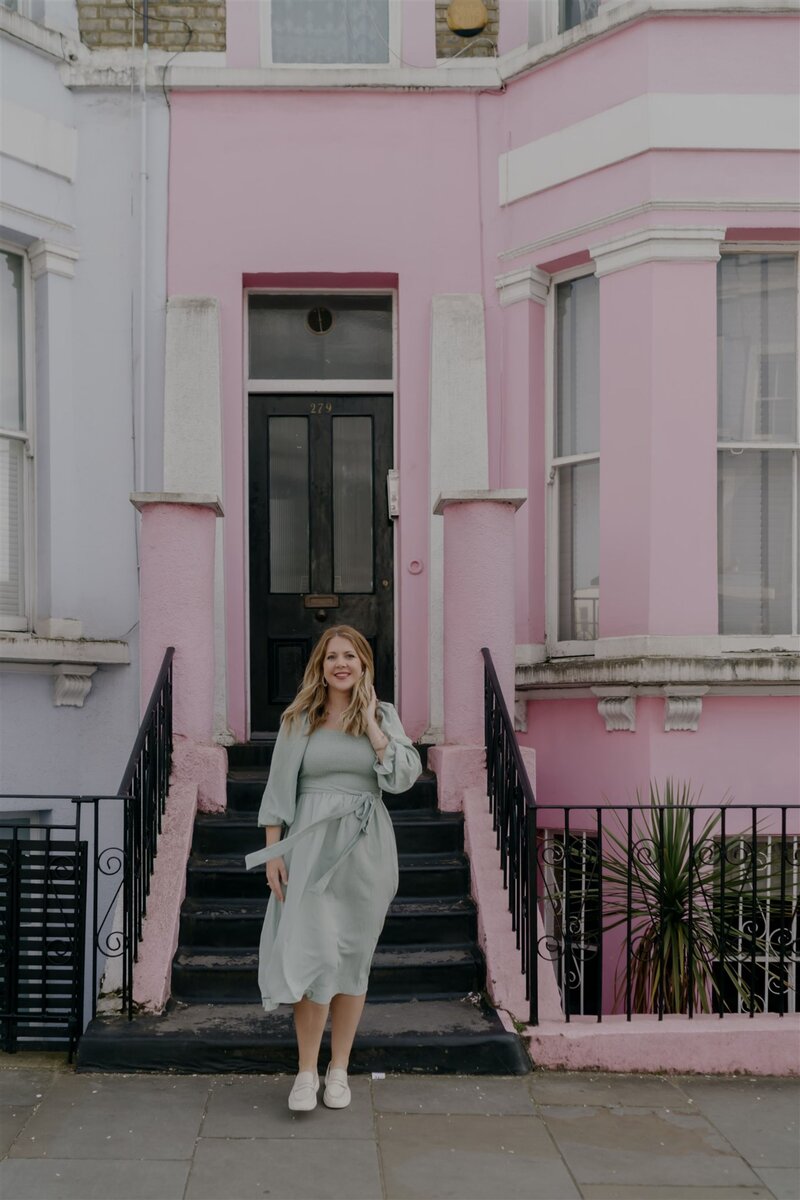 Woman in a soft green dress standing on steps in front of a pastel pink townhouse, smiling with one hand on the railing