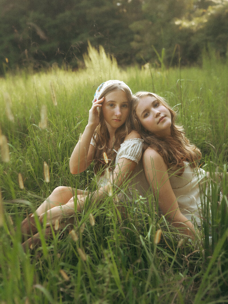 Two sisters in cream color outfits sit back to back in the tall lush green grass at Hyde Farm Park. 