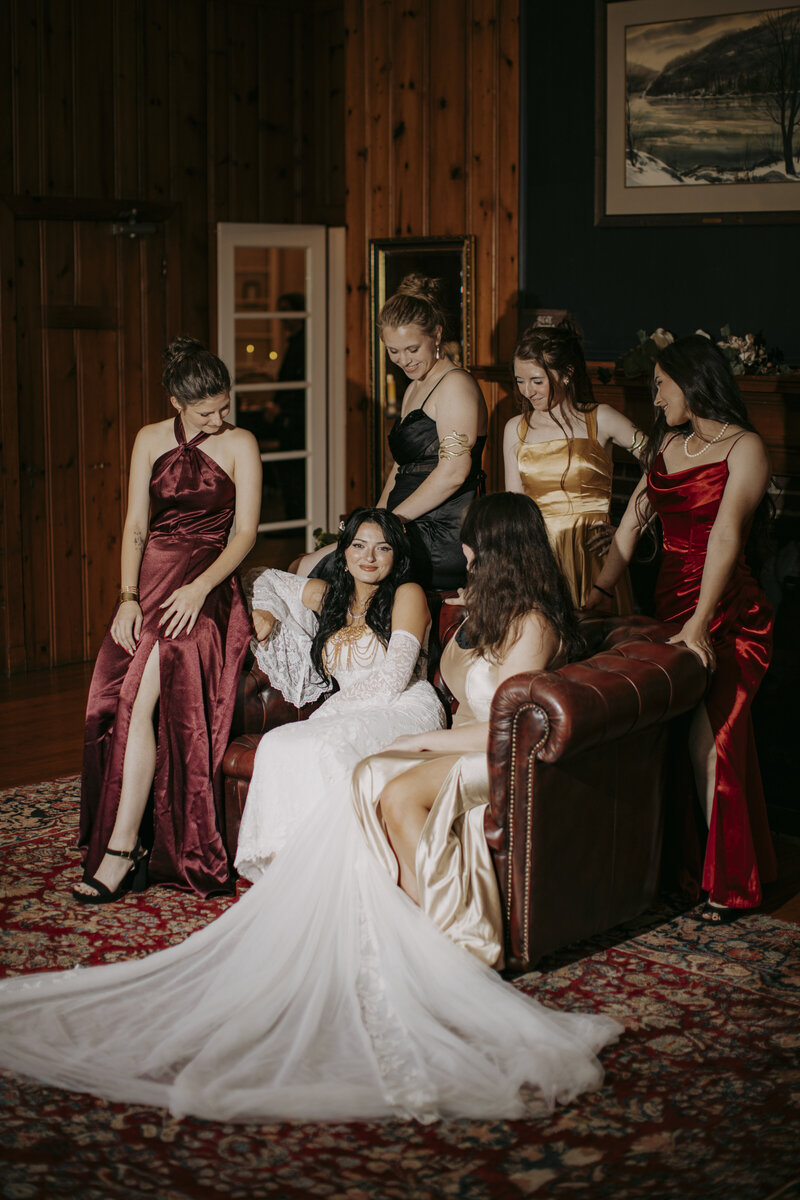 Bride surrounded by bridesmaids in rich burgundy dresses sharing a candid laugh inside a rustic bridal suite