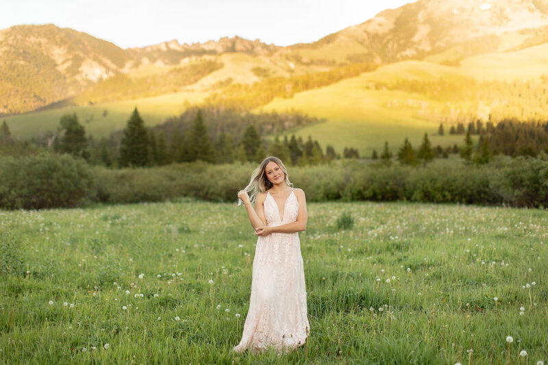 Touching the end of  a strand her hair, a young woman  smiles in front of a mountain.