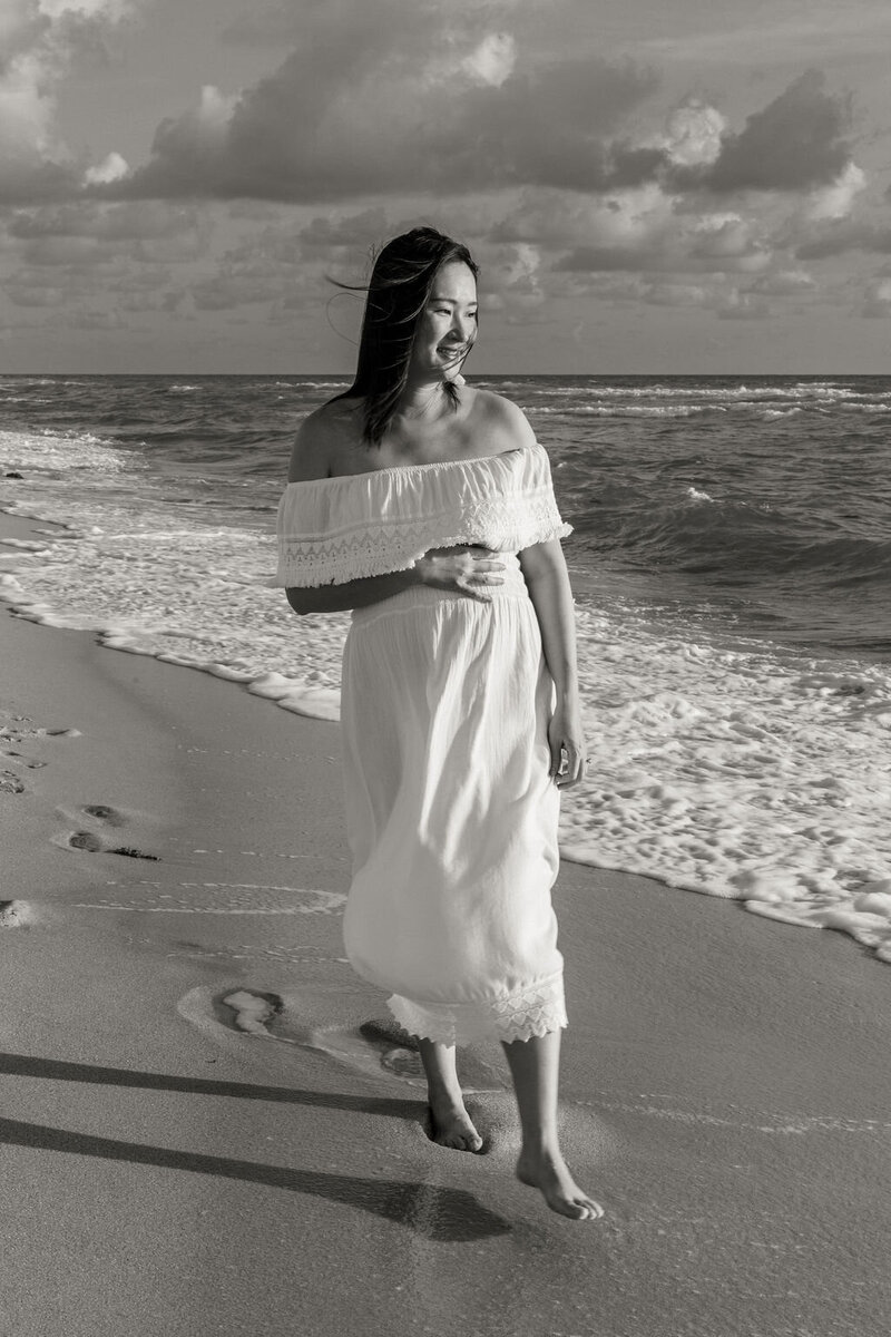 Pregnant woman in a white dress walking barefoot along the shore during a maternity session in Sunny Isles Beach.