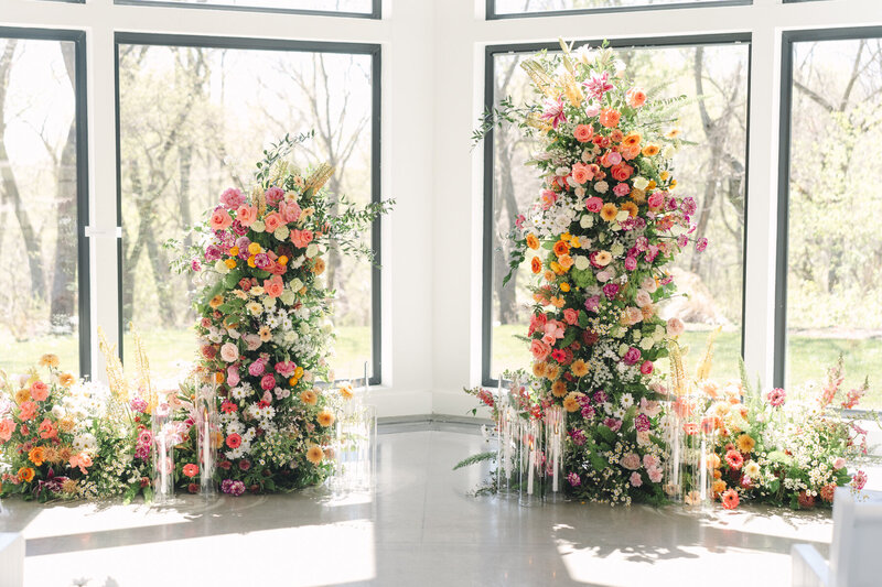 Luxury garden-style wedding ceremony installation featuring two tall, colorful floral pillars designed with roses, dahlias, ranunculus, daisies, greenery, and spring blooms, surrounded by ground arrangements and glass pillar candles inside a bright window-filled chapel—vibrant floral arch alternative for modern weddings.