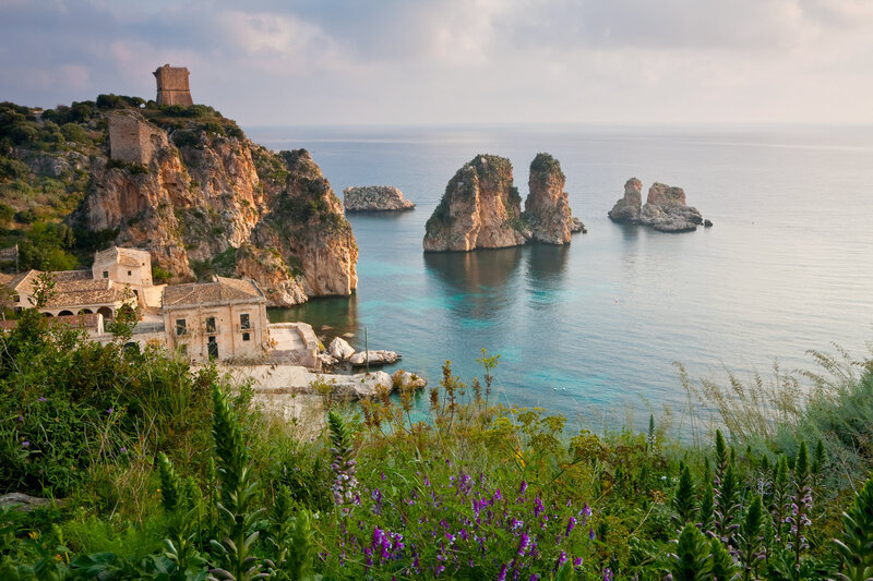 Coastal view of ancient buildings at Tonnara di Scopello in Sicily, Italy. Rugged cliffs and rock formations rise from the clear blue sea under a cloudy sky. Lush greenery and purple wildflowers line the foreground.