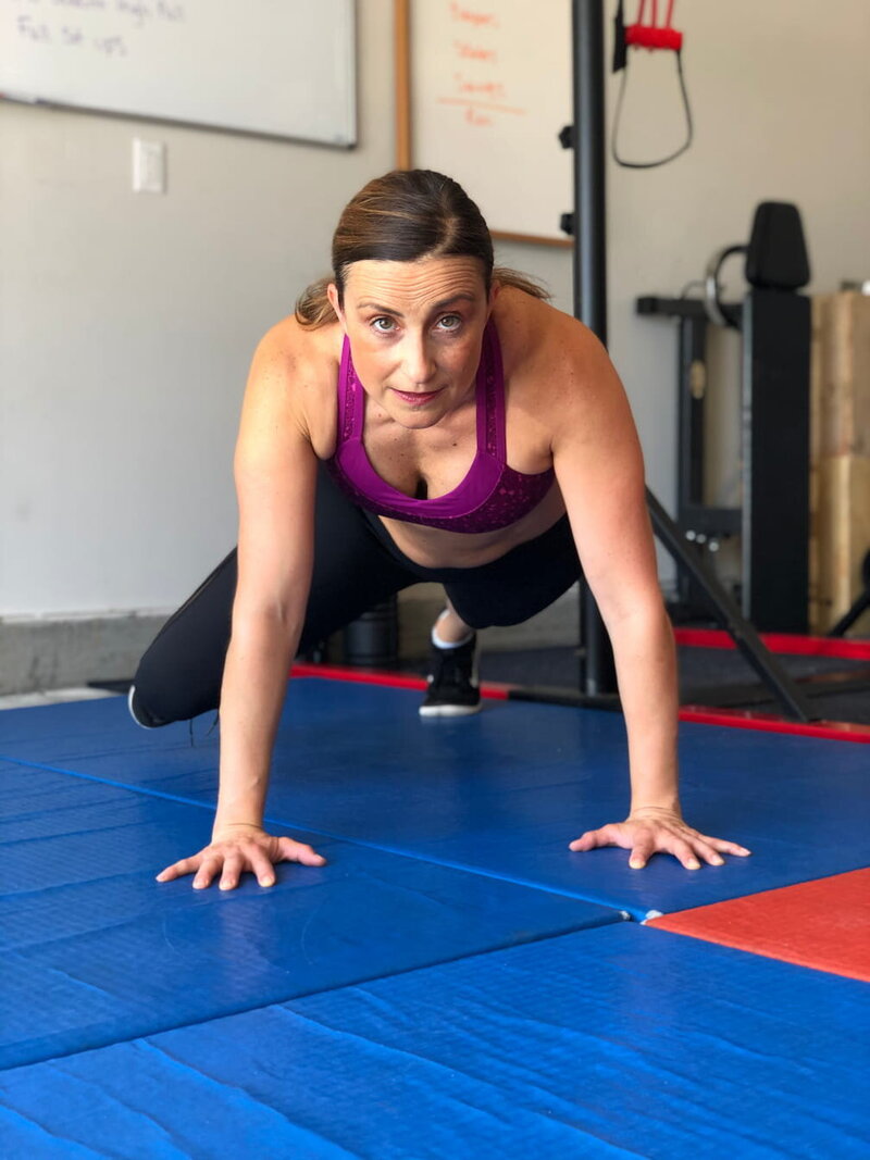 Vanessa Flores, a midlife strength coach, performing a plank-to-push-up variation on a blue mat in her home gym. She is focused, wearing a magenta sports bra and black leggings.