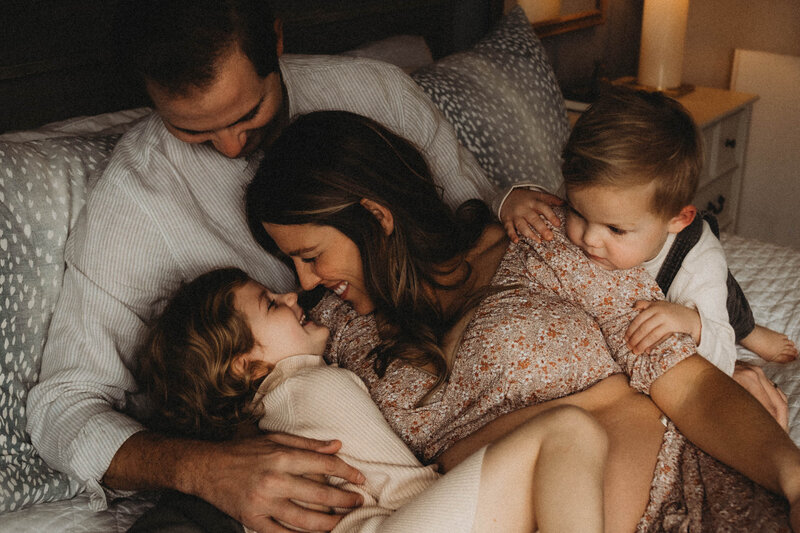 Relaxed in-home maternity photo of a mother-to-be sitting on a bed with husband and two children