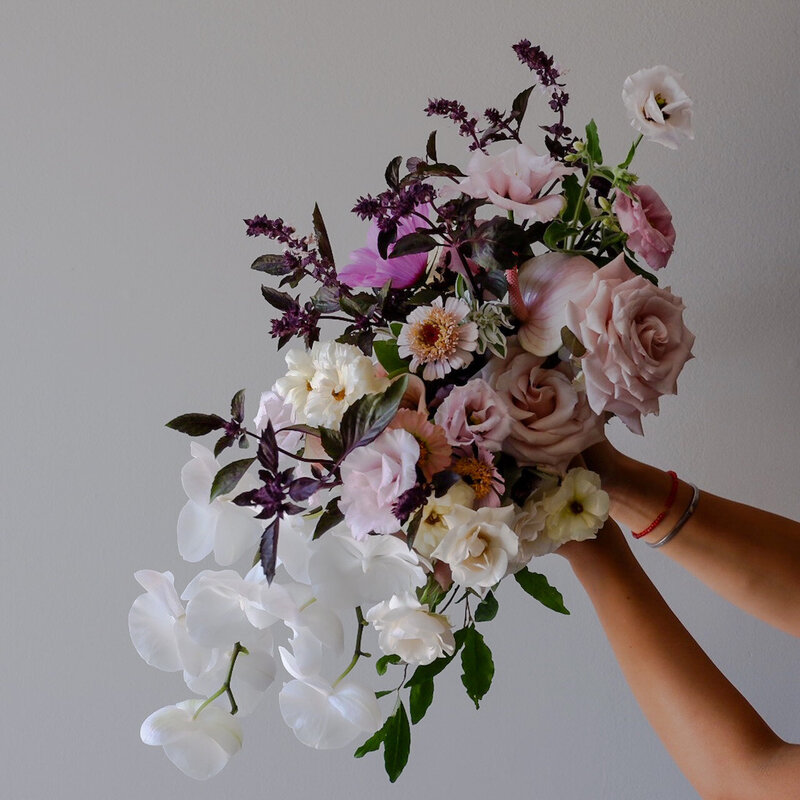 Lush bridal bouquet with white orchids, soft pink roses, deep purple foliage, and delicate accent flowers against a neutral background.