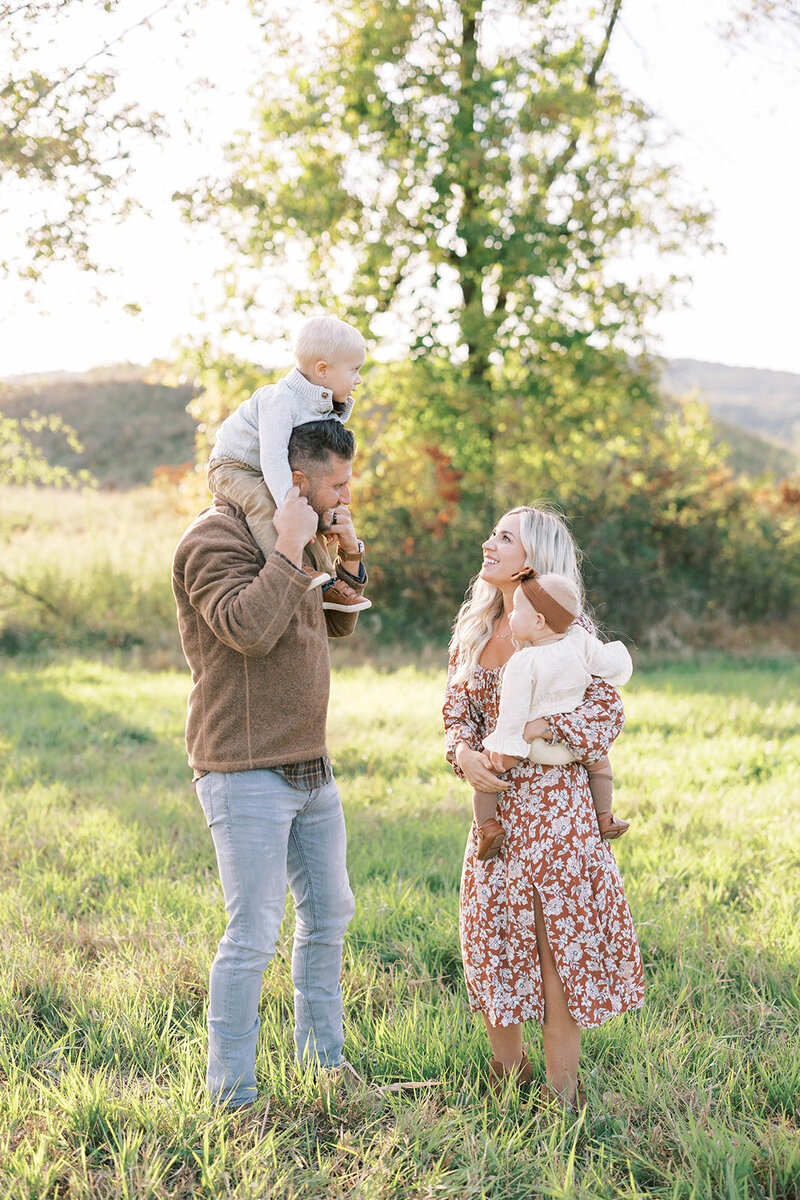 Dad holds toddler on his shoulders as mom holds baby in outdoor field at the base of the Appalachian mountains in Pennsylvanai