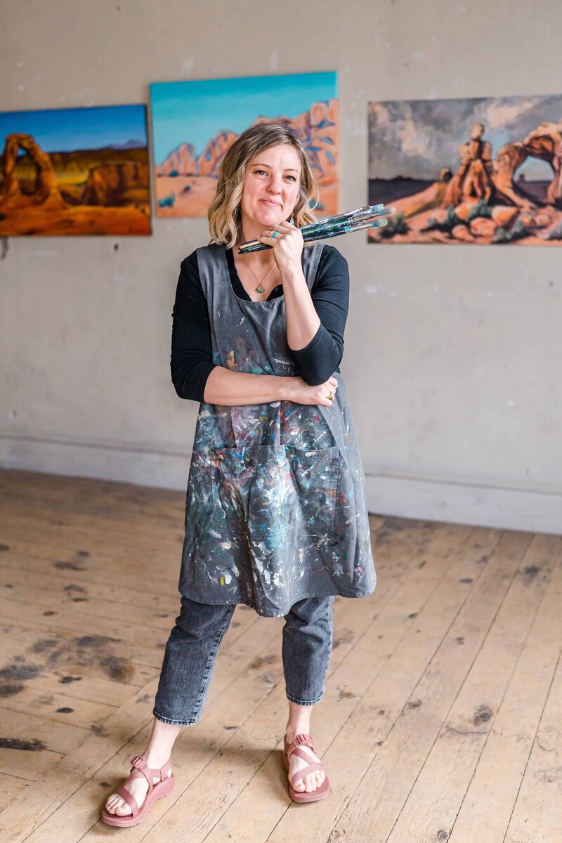An artist stands in her studio wearing a paint-splattered apron, holding brushes and smiling proudly in front of her landscape paintings.