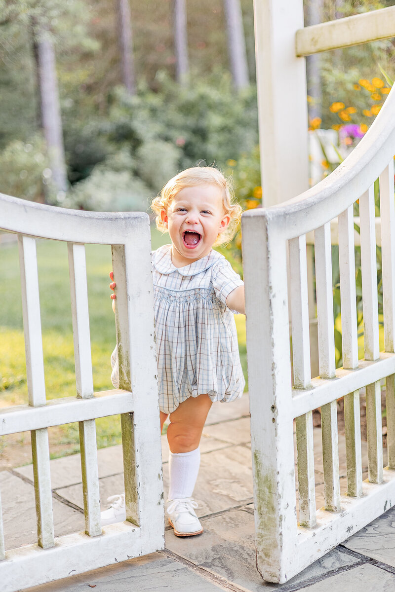 A smiling toddler in a pink dress and white bow holds onto a white garden gate.