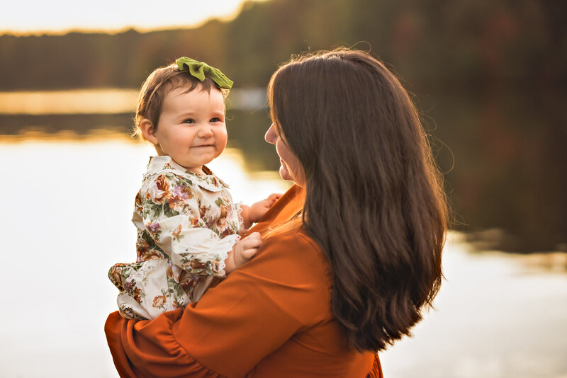 A mom in an orange dress is holding her baby at sunset.