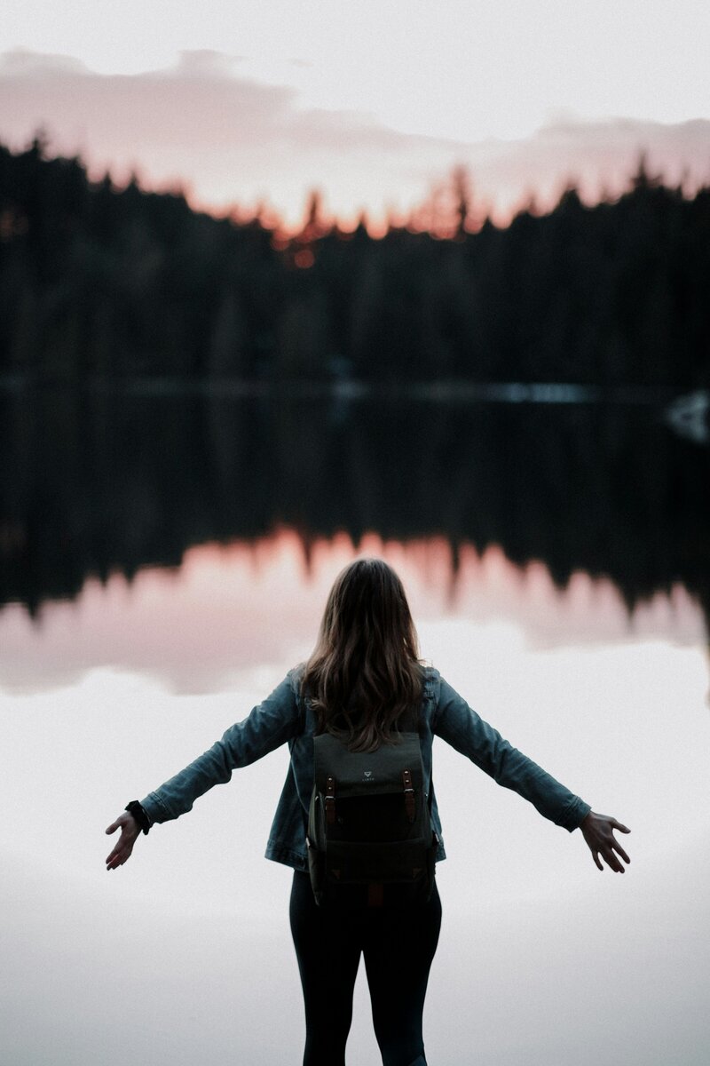 Aesthetic photo of a woman standing by a body of water surrounded by trees, representing peace, balance, and the holistic wellness approach of hormone therapy at Wellness & Hormone Health in Centennial, Colorado.