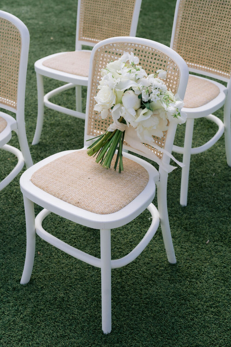 Close-up of a white bridal bouquet resting on a white cane-back chair on grass at an outdoor ceremony.