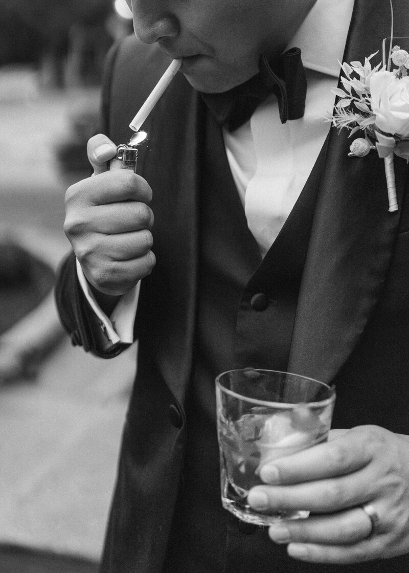 A groom lights a cigarette, whiskey glass in hand.